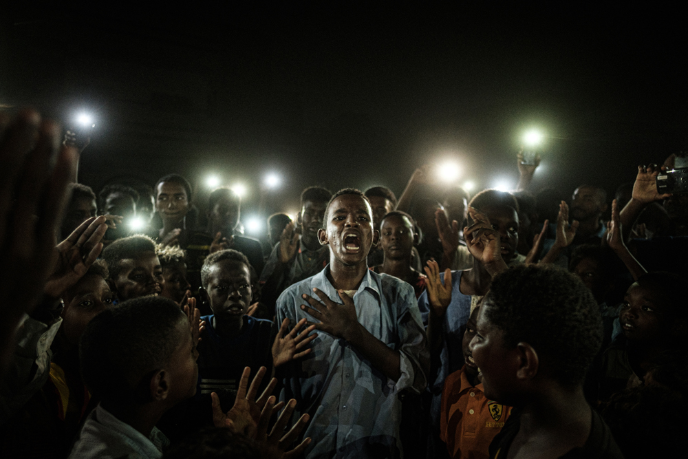People chant slogans as a young man recites a poem, illuminated by mobile phones, before the opposition's direct dialog with people in Khartoum on June 19, 2019. - People chanted slogans including "revolution" and "civil" as the young man recited a poem about revolution. (Photo by Yasuyoshi CHIBA / AFP)